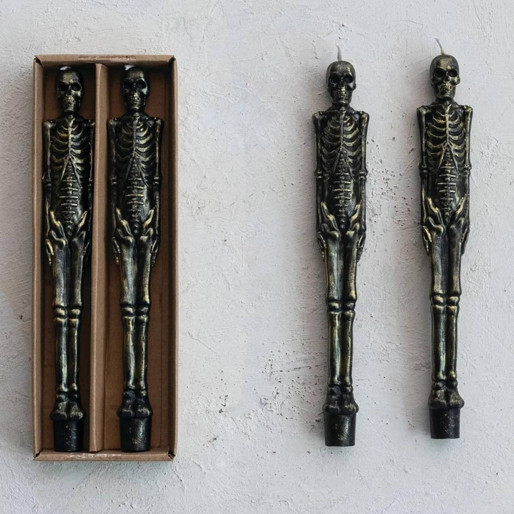 Two skeleton taper candles on a textured white surface with two in a box beside.
