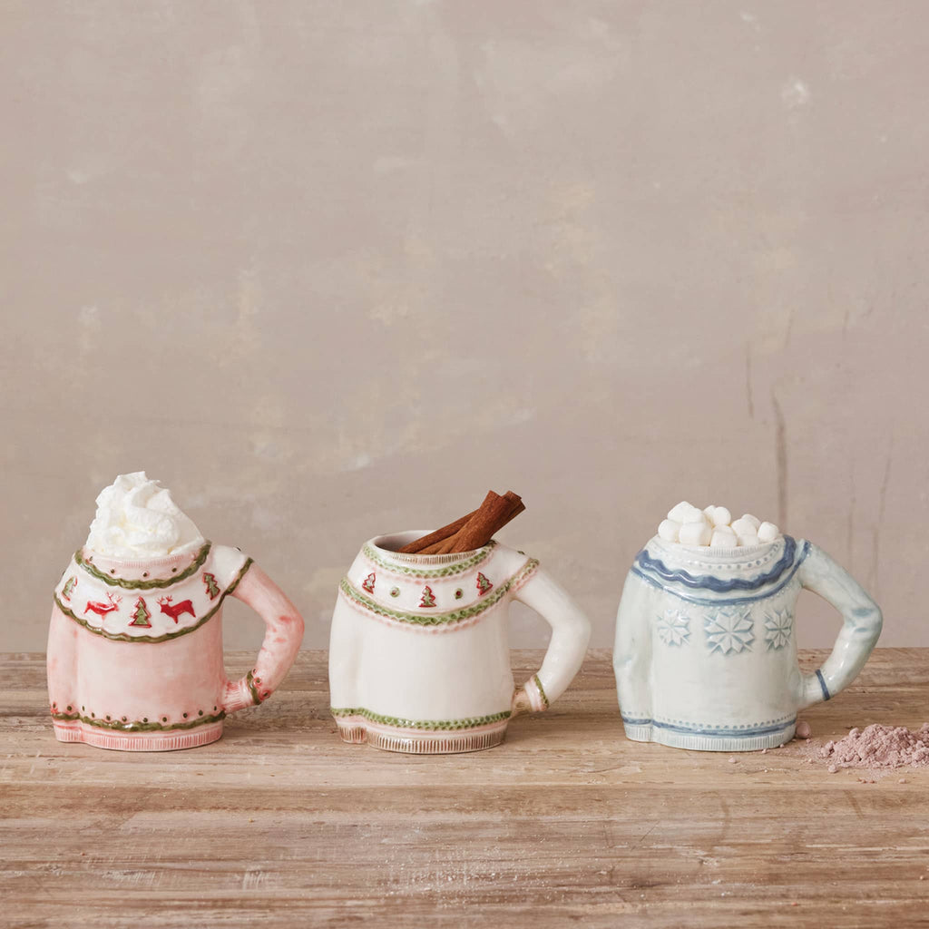 Three ceramic hot chocolate mugs with sweater designs on a wooden surface.