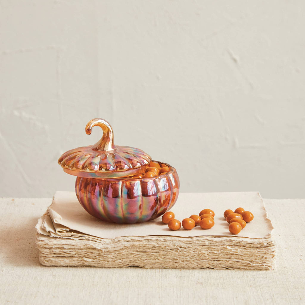 Creative Co-op Recycled Glass Pumpkin-Shaped jar with iridescent finish and lid with gold tone stem handle. Filled with candies on stack of tan paper with tan background.