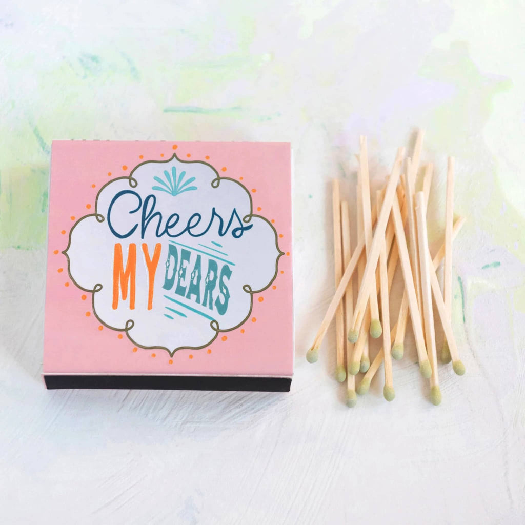 Creative Co-op pink box of wood safety matches with "Cheers My Dears" on the front, with a pile of green-tipped matches beside it.