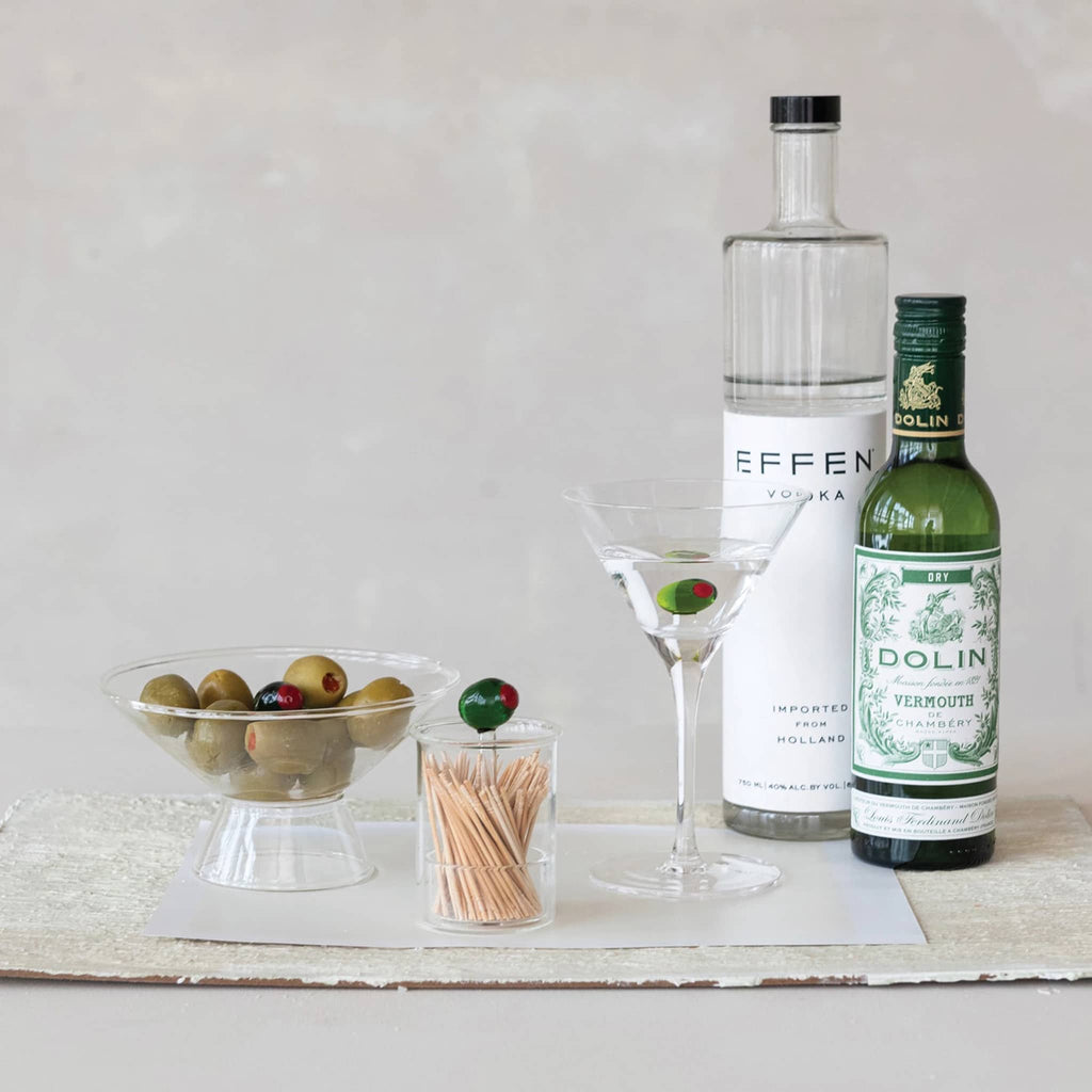 Clear glass footed serving bowl with a glass green olive in middle, with olives and matching martini glass with vermouth and vodka bottles.