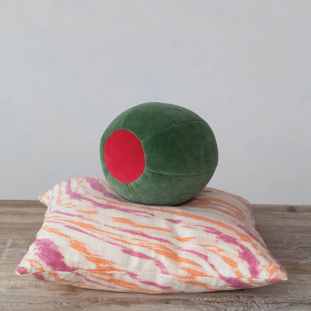 Green olive shaped cotton velvet pillow with red center, front angle, on other throw pillow with orange and pink design.