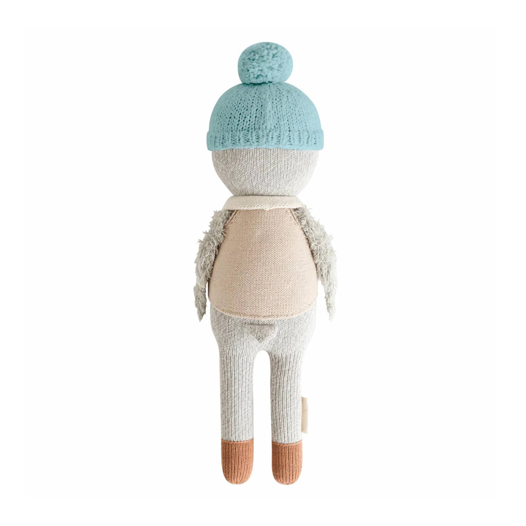 Stuffed knit toy penguin wearing a blue knitted hat, beige sweater, and white pants, back view, on a white background.