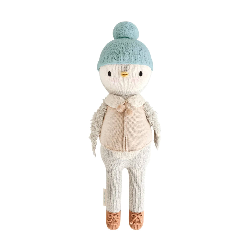 Stuffed knit toy penguin wearing a blue knitted hat, beige sweater, and white pants on a white background.