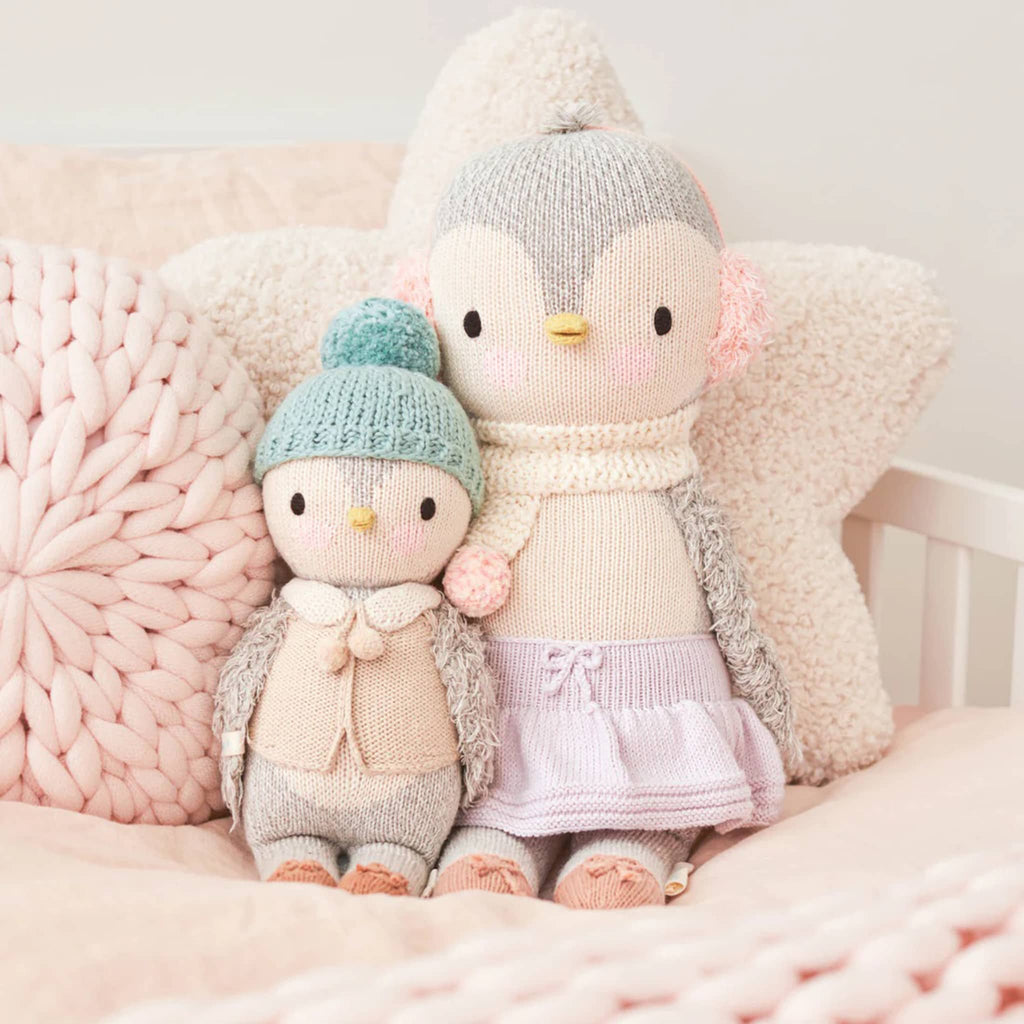 Two plush penguin toys wearing knitted hats and sweaters on a soft pink background.