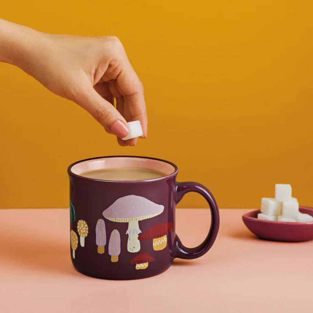 Person adding sugar to a mug of tea with a mushroom design on an orange background.