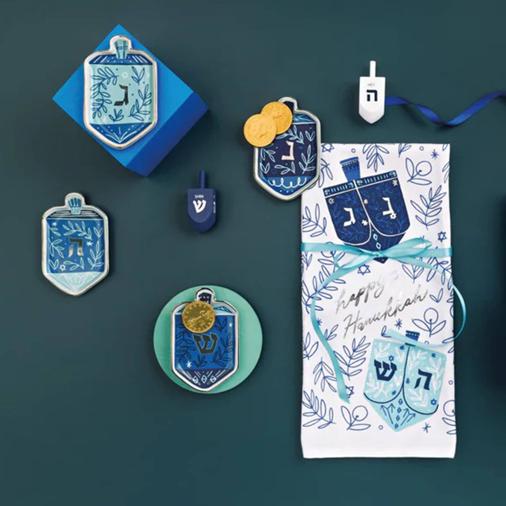 Collection of Hanukkah-themed decorative items including dreidel dishes and a dishtowel with dreidels and menorahs, on a dark green background.