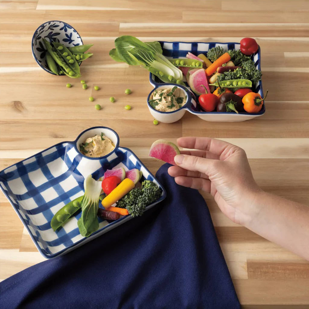 Hand holding a vegetable over a dish with built in dip bowl with various vegetables and a small bowl of dipping sauce on a wooden surface.