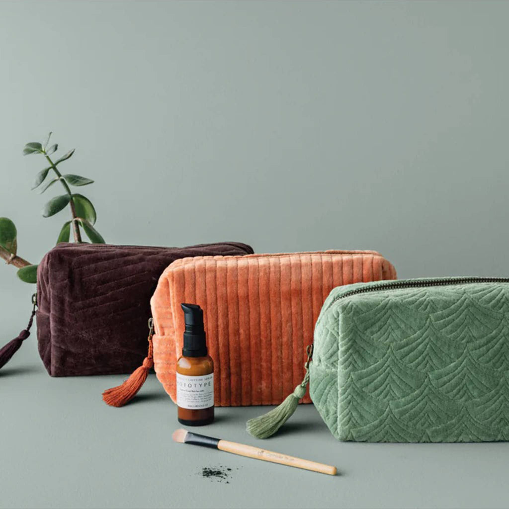 Three textured cosmetic bags in fig, copper, and jade on a sage green surface with a plant in the background.