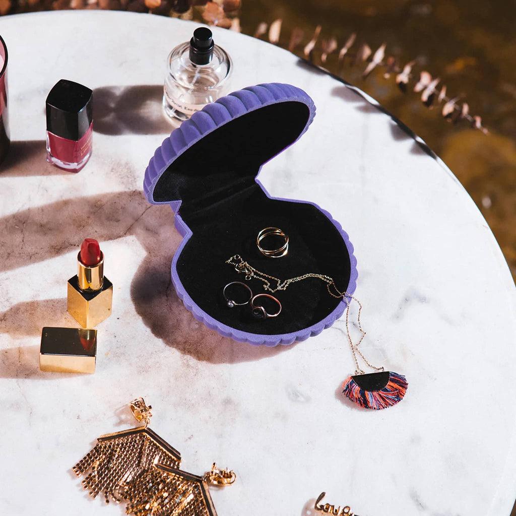 Purple shell-shaped jewelry box with jewelry on a marble surface with lipstick and nail polish.