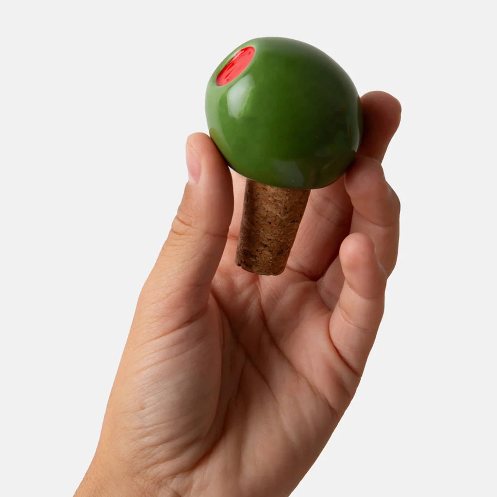 Hand holding a ceramic green olive bottle stopper with cork base on a white background.