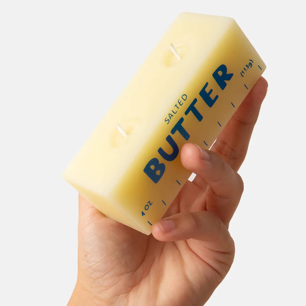 Hand holding a rectangular yellow paraffin and beeswax candle that looks like a block of butter on a white background.