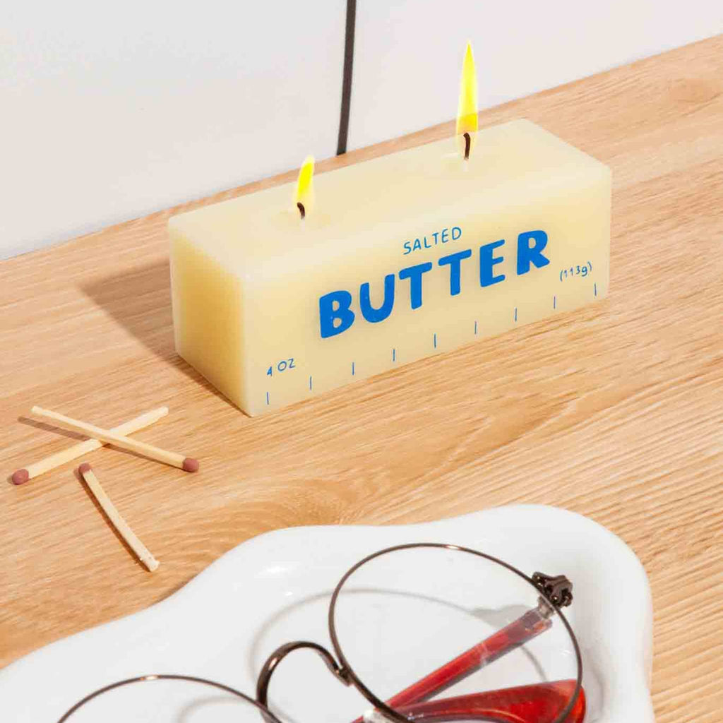 Butter-shaped candle labeled 'Salted Butter' on a wooden surface with matches and glasses.
