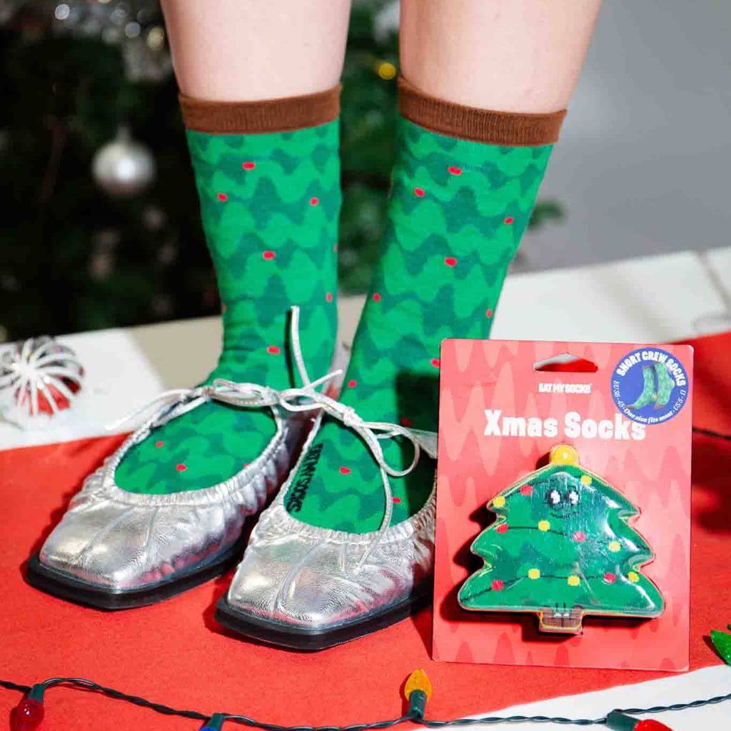 Green Christmas tree-patterned socks with red dots, silver shoes, and a packaging card on a festive background.
