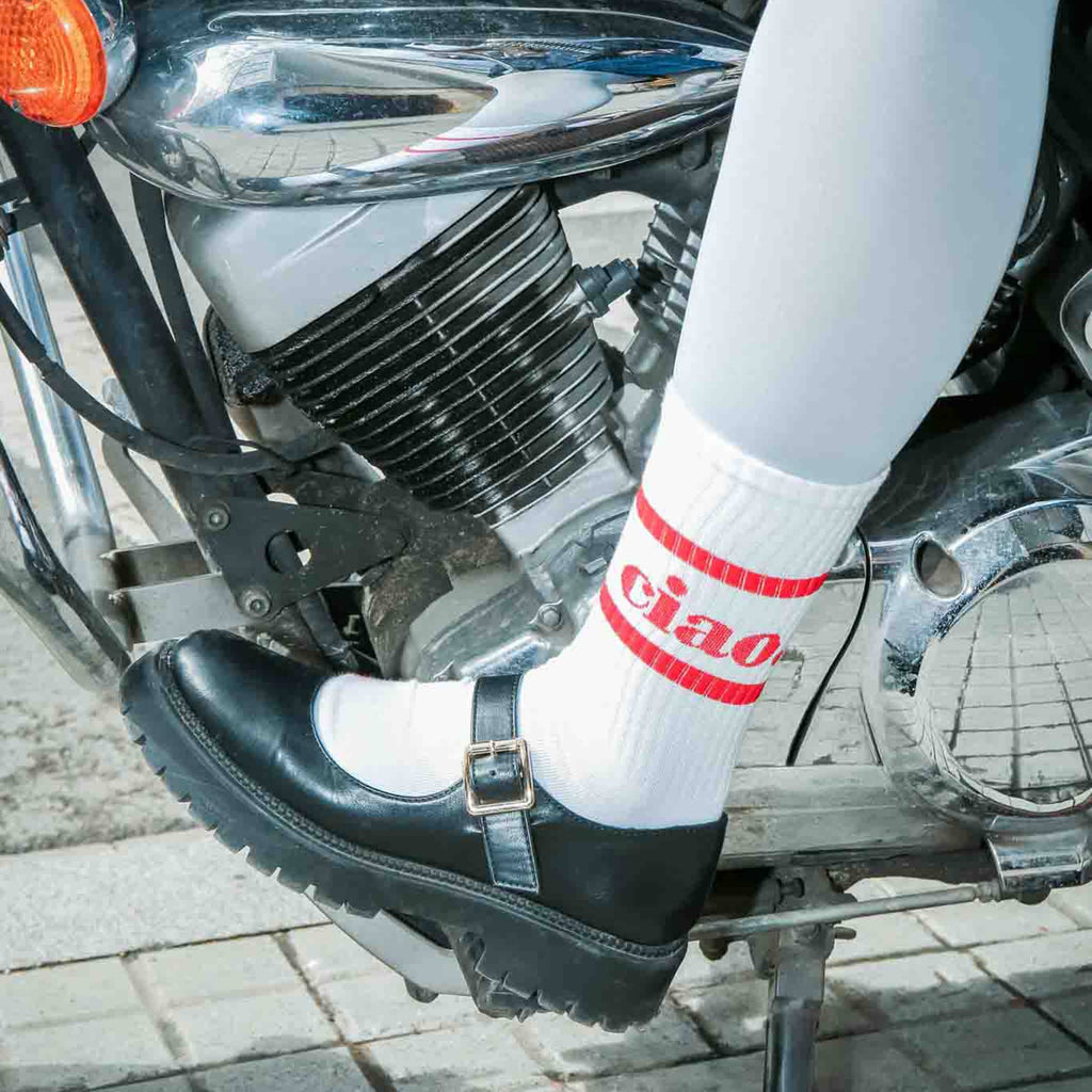 Person wearing a black shoe and white sock with red "ciao" text on a motorcycle.