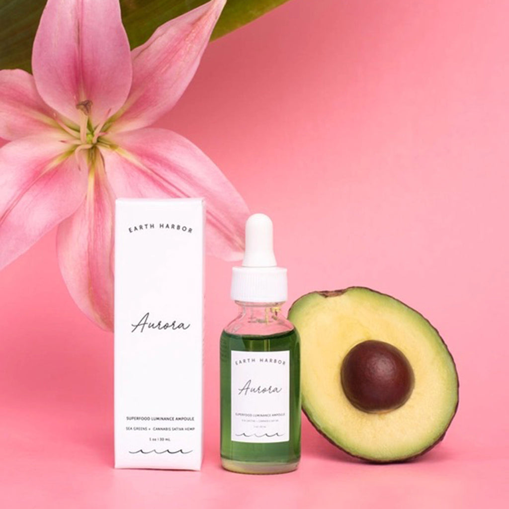Earth Harbor Aurora Superfood Luminance Ampoule in 1 ounce clear glass bottle with eyedropper, front view, with white box packaging, an avocado and a tropical flower on a pink background.