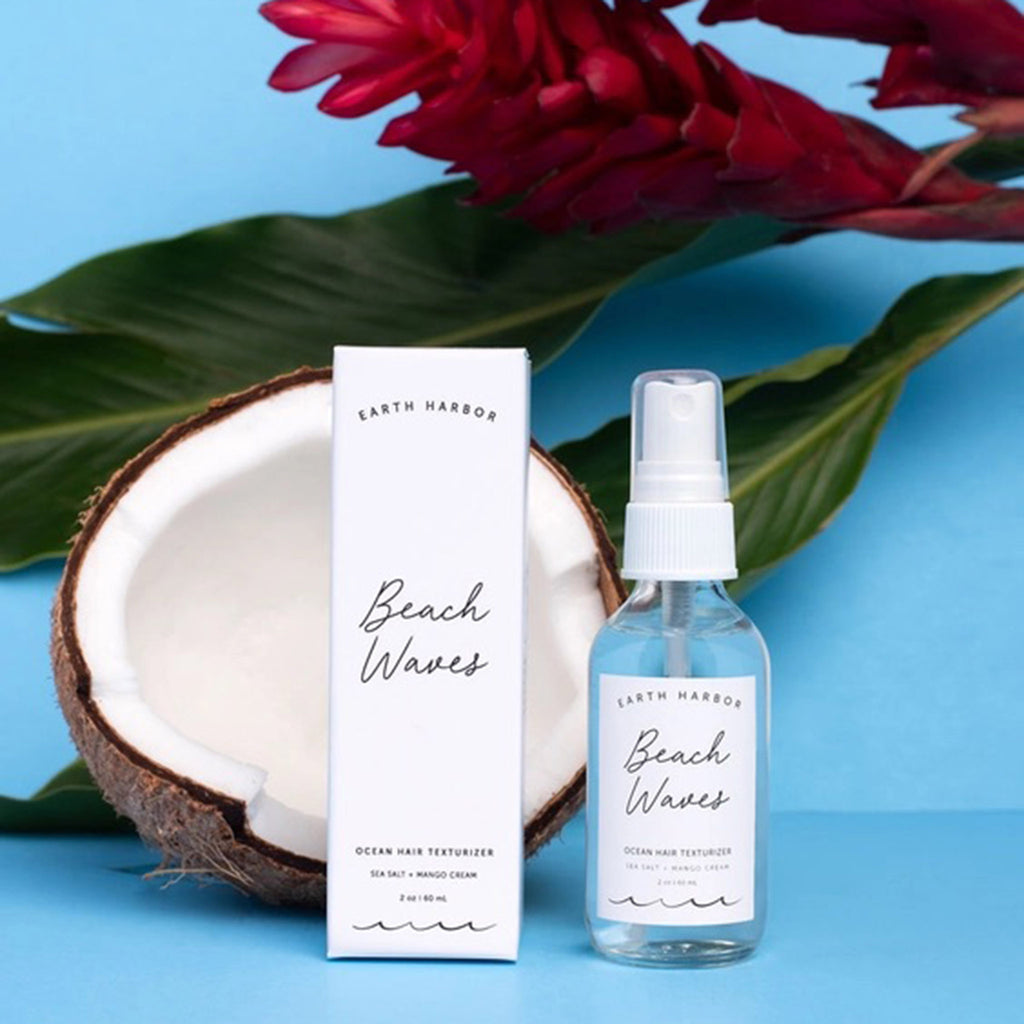Earth Harbor Beach Waves Ocean Hair Texturizer in clear glass spray bottle, front view with white box packaging and a coconut half and tropical flower on a blue background.