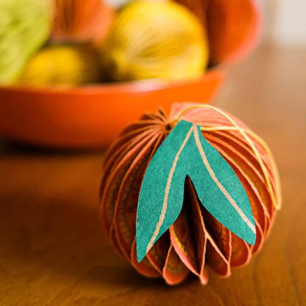 Decorative orange-shaped honeycomb paper ornament with a leaf on wooden surface.