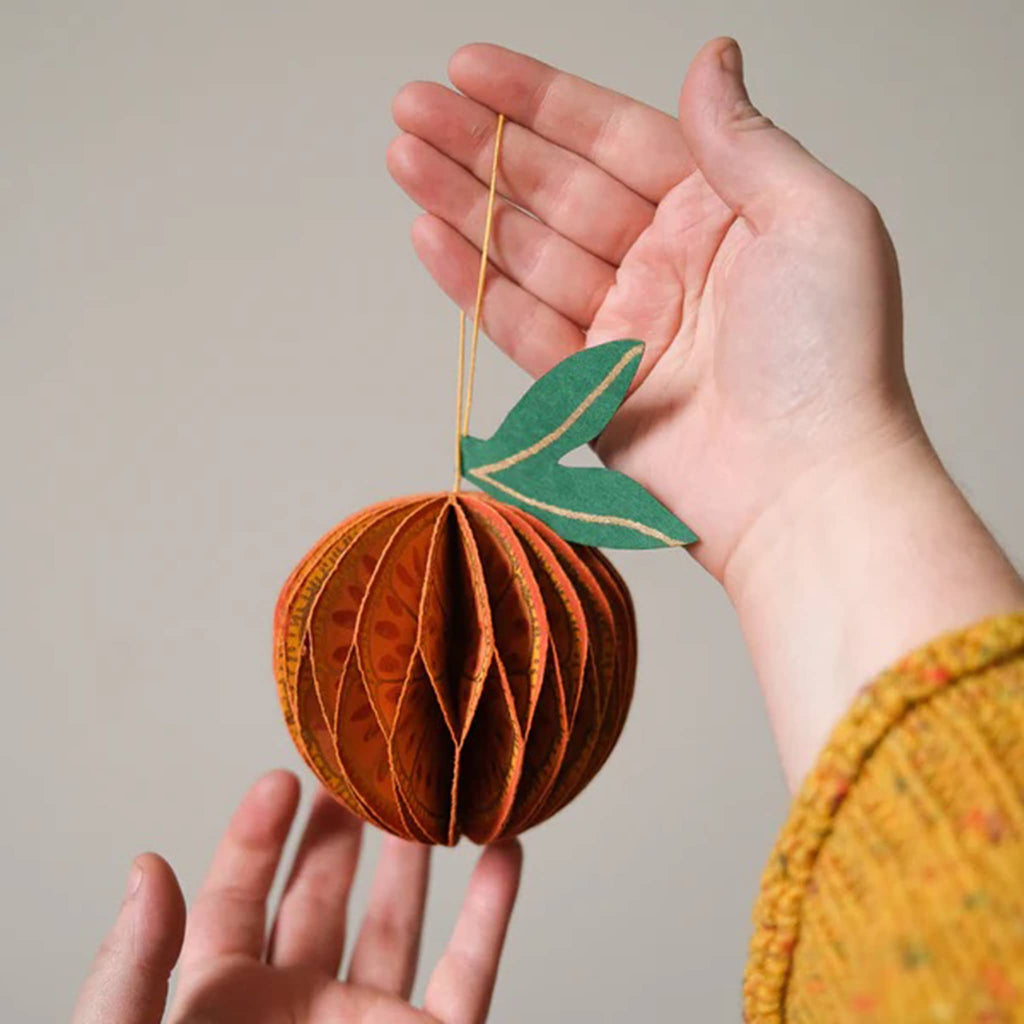 Hand holding a paper ornament shaped like an orange with a green leaf against a neutral background.