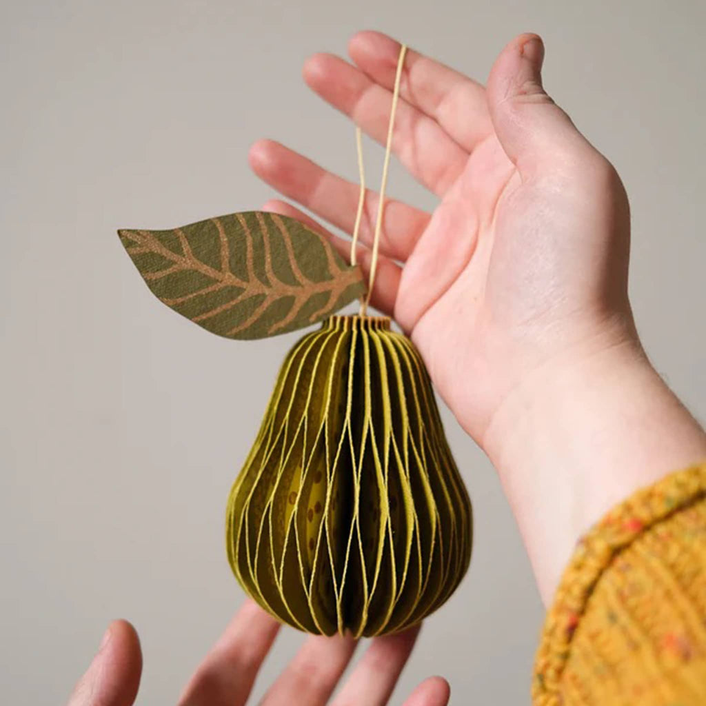Hand holding a decorative paper pear with a leaf against a neutral background.
