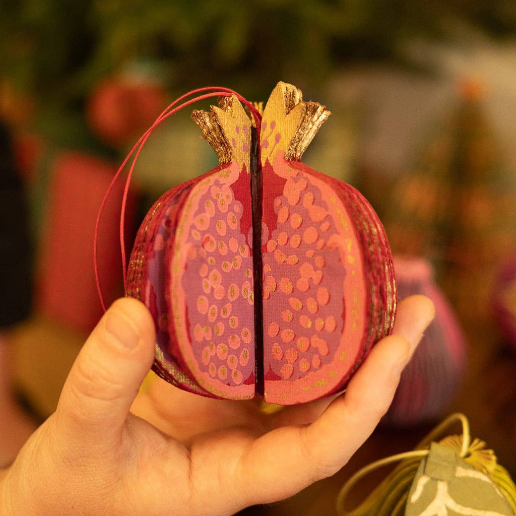 Hand holding a decorative pomegranate ornament with a blurred Christmas tree background.