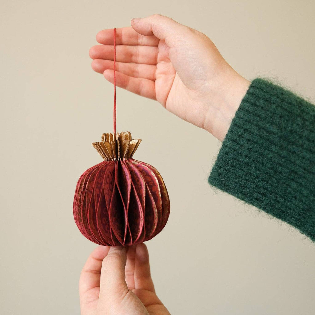 Hand holding a decorative red pomegranate-shaped honeycomb paper ornament against a neutral background.