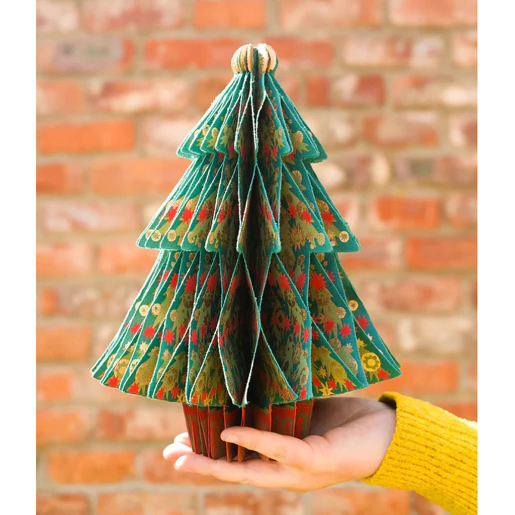 Hand holding a decorative paper Christmas tree against a brick wall.