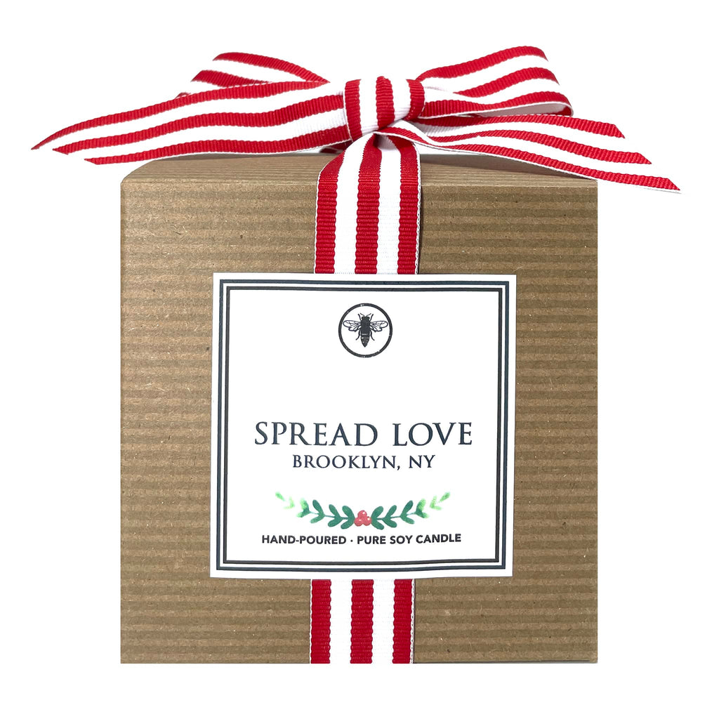 Ella B Candles Spread Love leather, tea and pomegranate scented soy wax candle in kraft box packaging with red and white striped grosgrain bow, front view.