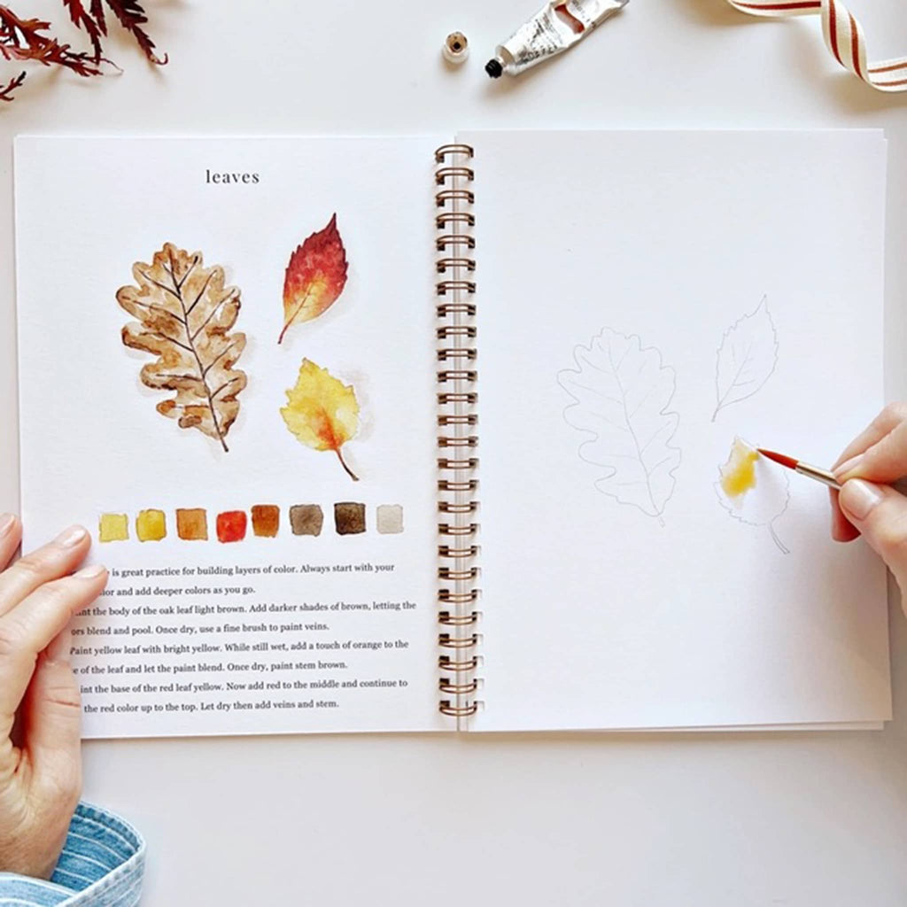 Open book with leaf illustrations and color swatches, held by a person with a paintbrush.
