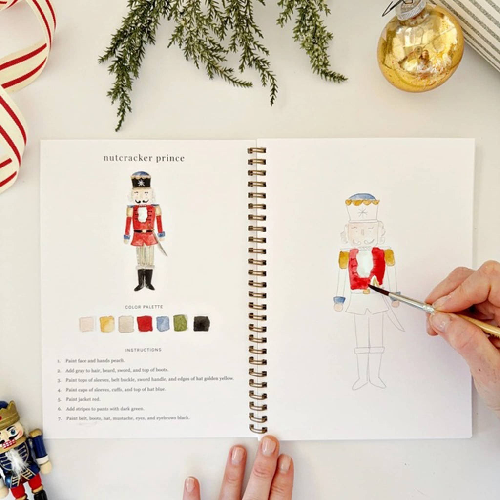 Person painting a nutcracker illustration in a book with Christmas decorations around.
