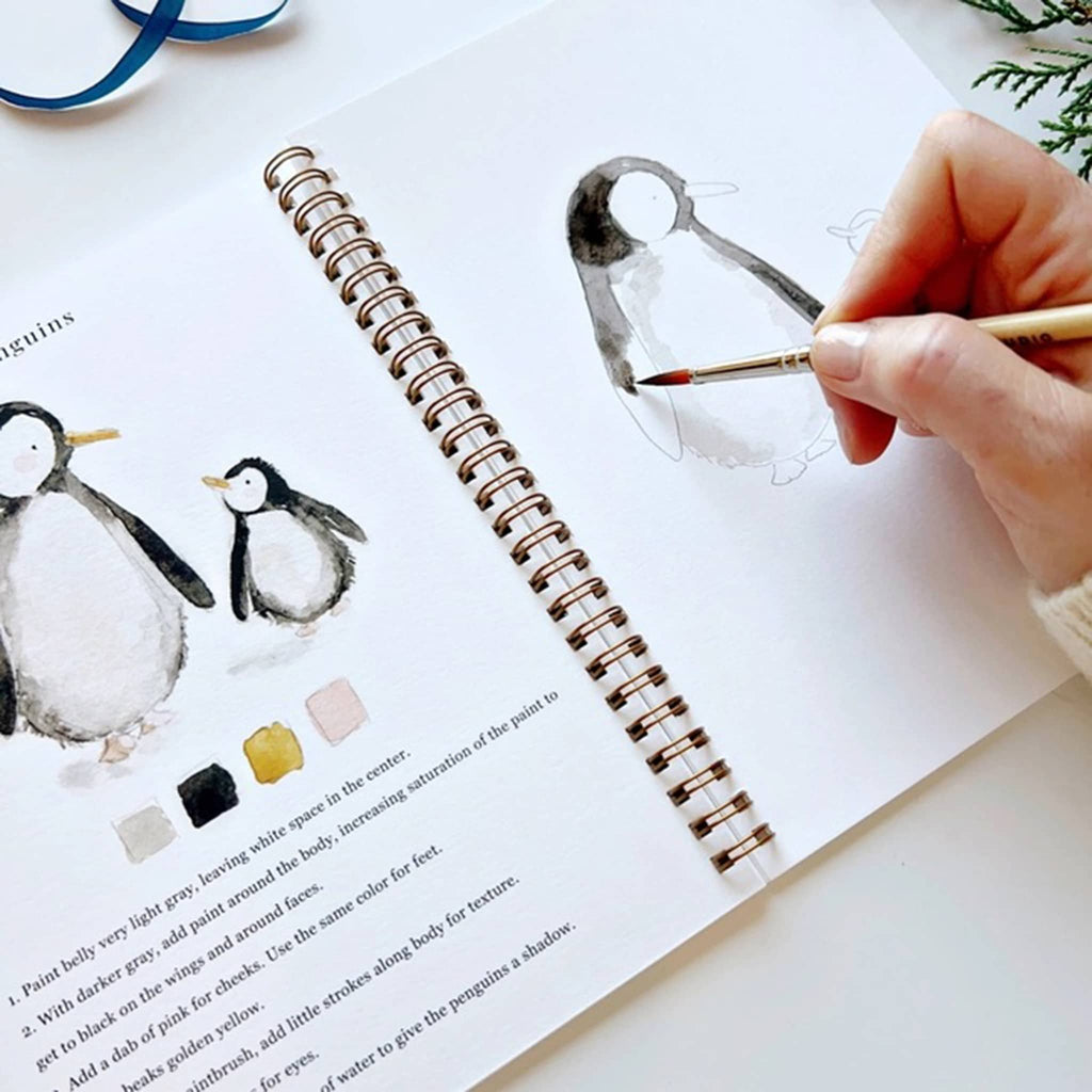 Person painting a penguin illustration in a sketchbook with watercolors.