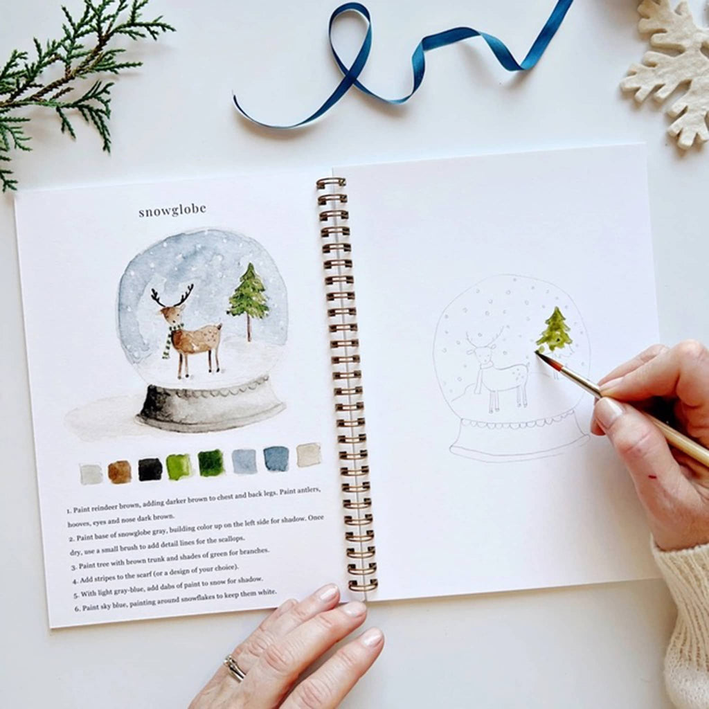 Person painting a snow globe illustration in a book with a paintbrush.
