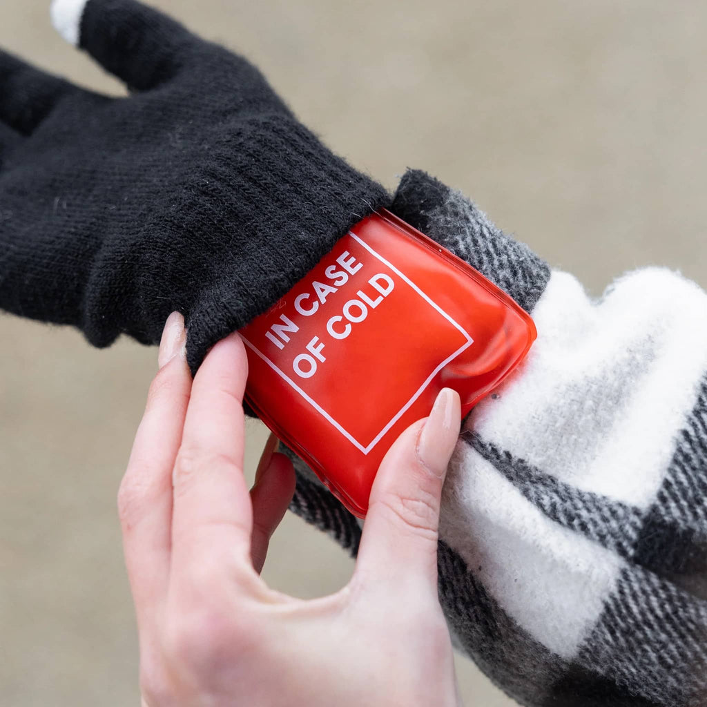 Hand sliding a red hand warmer with 'In Case of Cold' under a black glove on a neutral background.