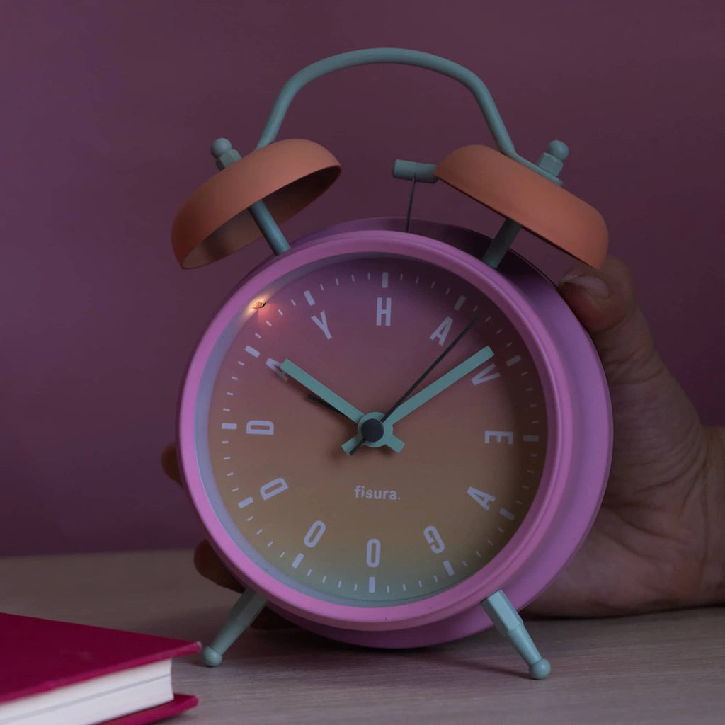 Pink alarm clock with light on, held by a hand against a purple background.
