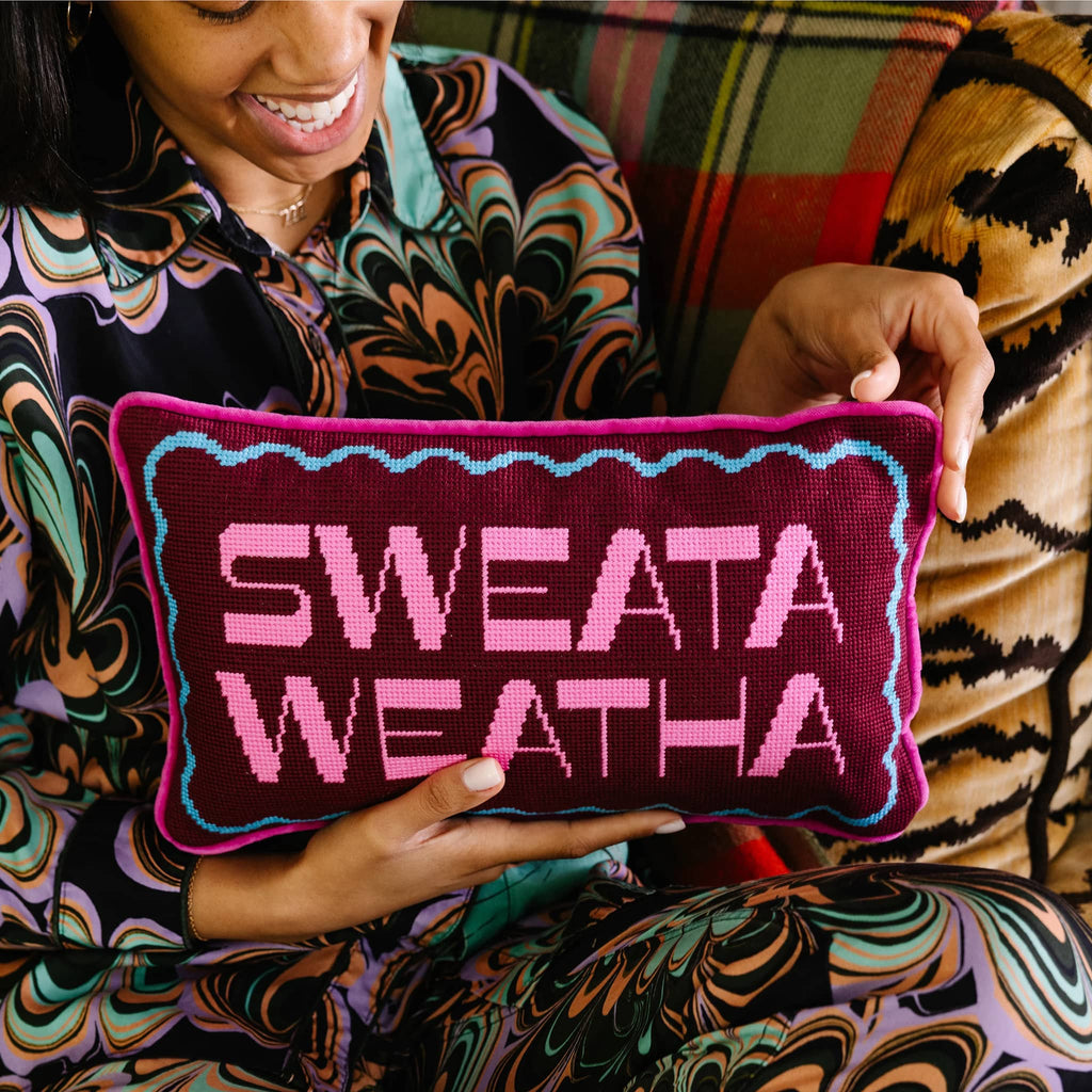 Person holding a pillow with 'SWEATA WEATHA' text in a colorful pattern.