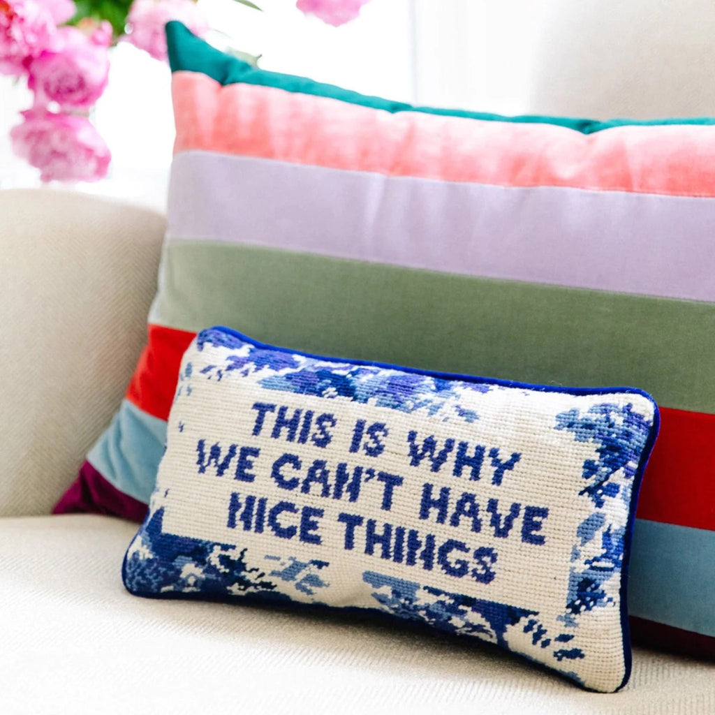 Furbish studio needlepoint pillow, cream with blue floral stitching and "this is why we can't have nice things" on the front on a couch in front of a velvet colorful striped pillow.