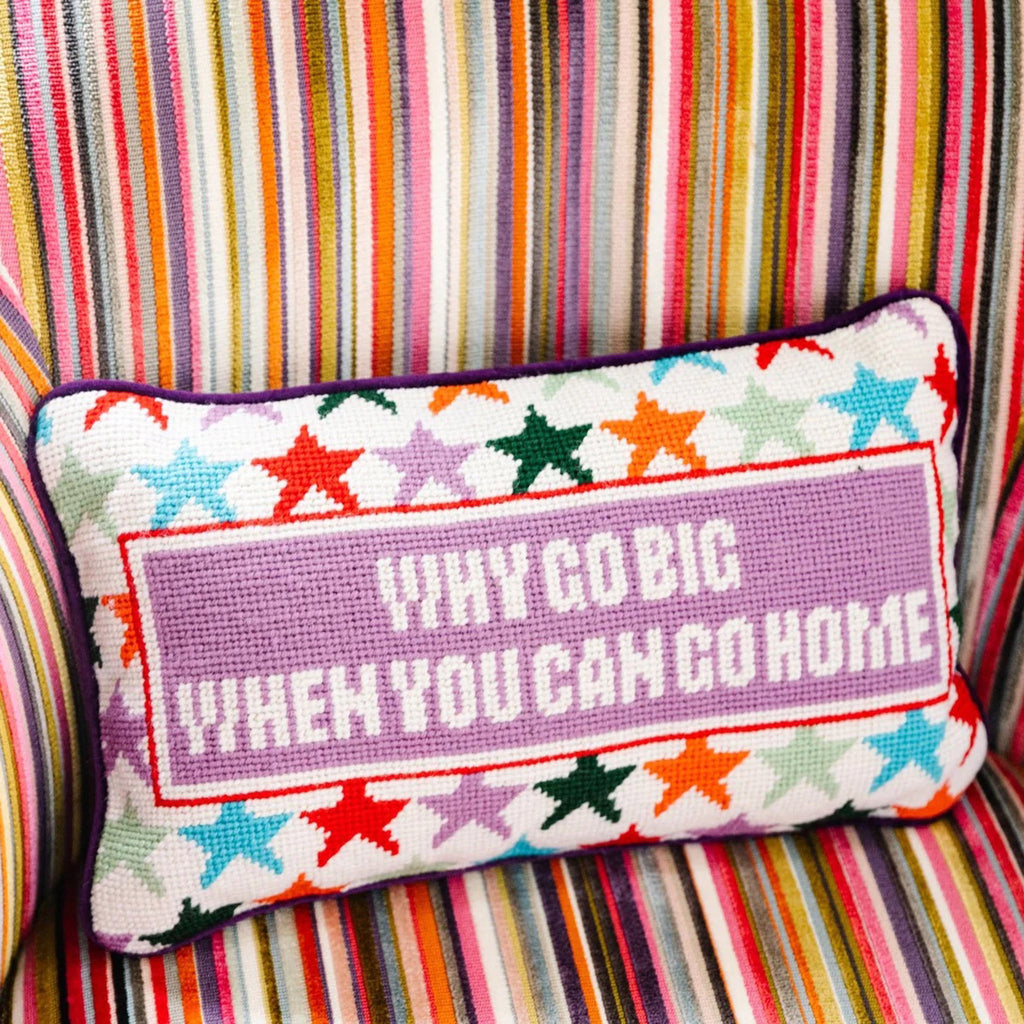 Furbish studio needlepoint pillow, white with colorful stars and "why go big when you can go home" on the front, sitting in a colorful striped chair.