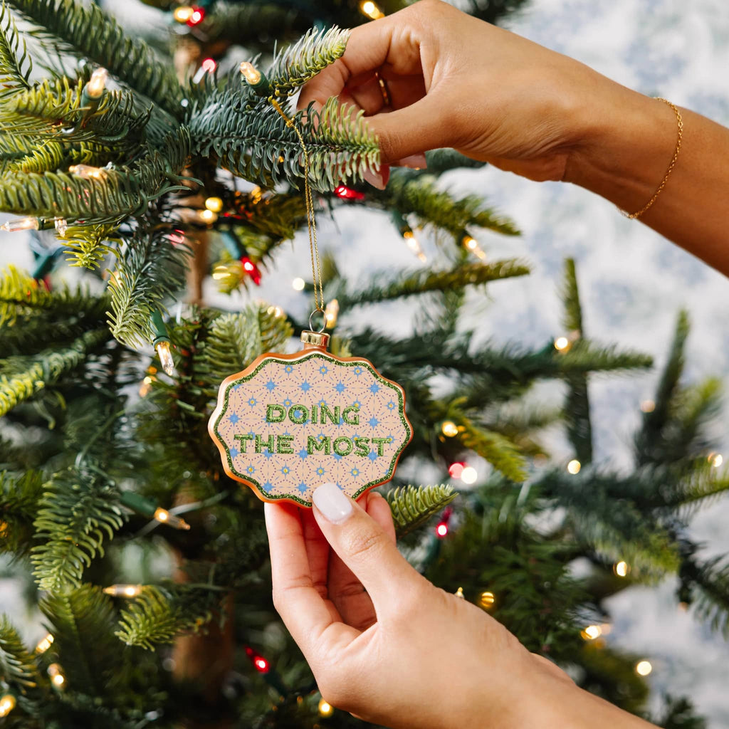 Hand holding a Christmas ornament with 'Doing the Most' text against a decorated tree background.