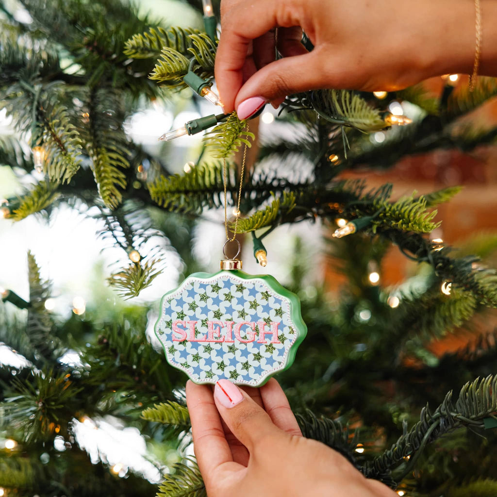 Hand holding a Christmas ornament with 'SLEIGH' text against a decorated tree background.