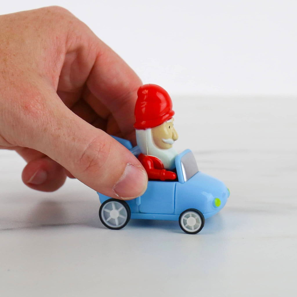 Toy figure of a gnome in a red hat inside a blue car held by a hand on a white background.
