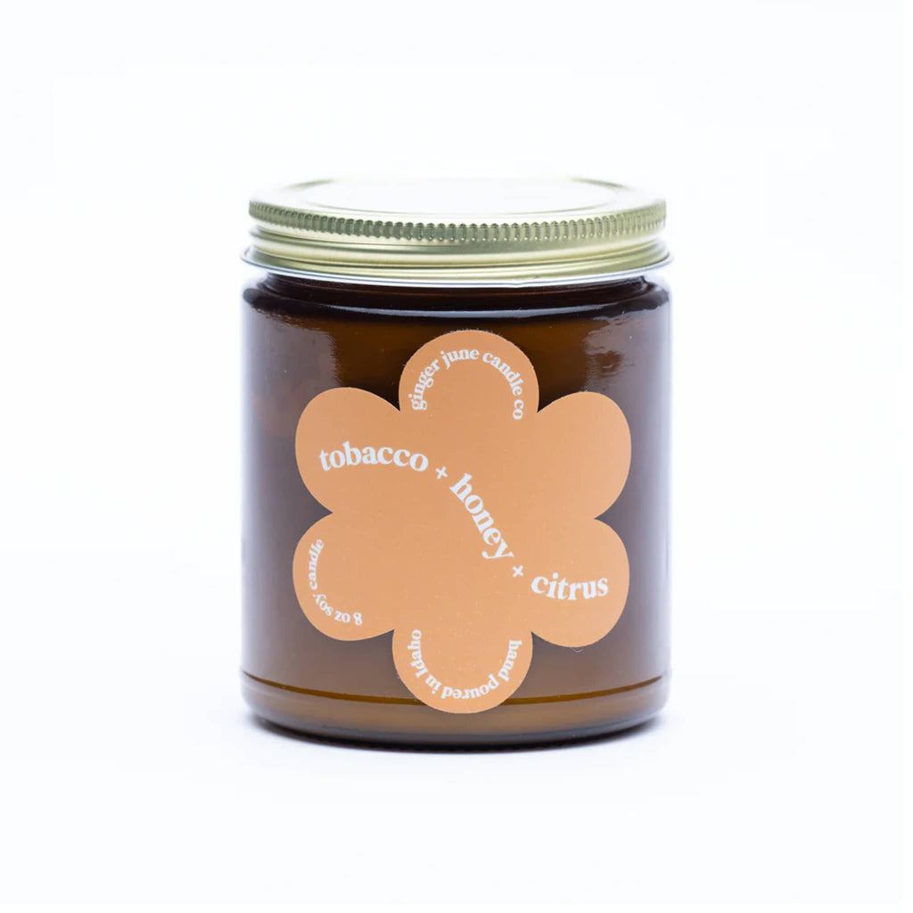 Ginger June 9 ounce Tobacco, Honey and Citrus scented soy wax candle in amber jar with gold screw-on lid and daisy-shaped orange label.