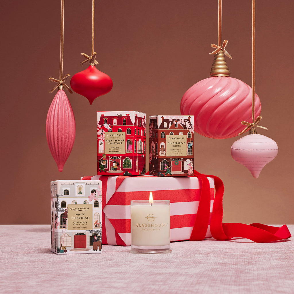 Glasshouse Fragrances Gingerbread House, Night Before Christmas and White Christmas scented holiday votive candles in a group shot with a lit votive, a red and pink striped gift and hanging vintage style ornaments on a pink background.
