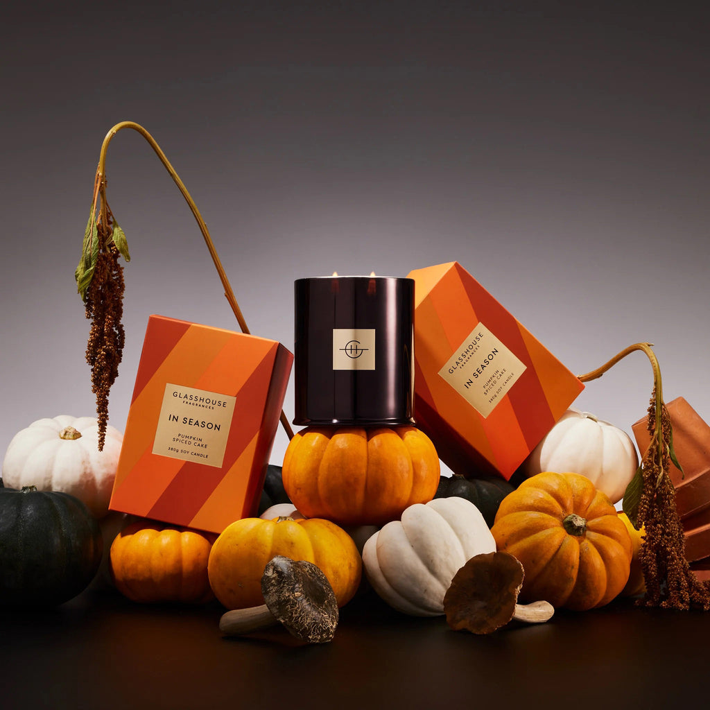 Glasshouse Fragrances In Season Pumpkin Spiced Cake scented soy wax candle in glossy black glass jar, shown with wicks lit and 2 gift boxes leaning against it on a pile of mini pumpkins.