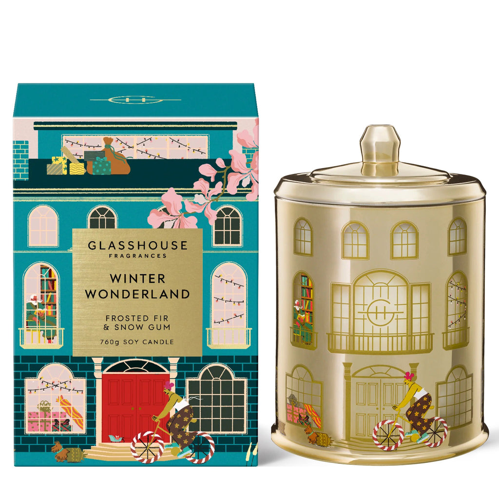 Glasshouse Fragrances Winter Wonderland 26.8 ounce frosted fir and snow gum scented soy wax candle in house illustrated gold glass jar with lid beside a blue and pink house gift box, front view.