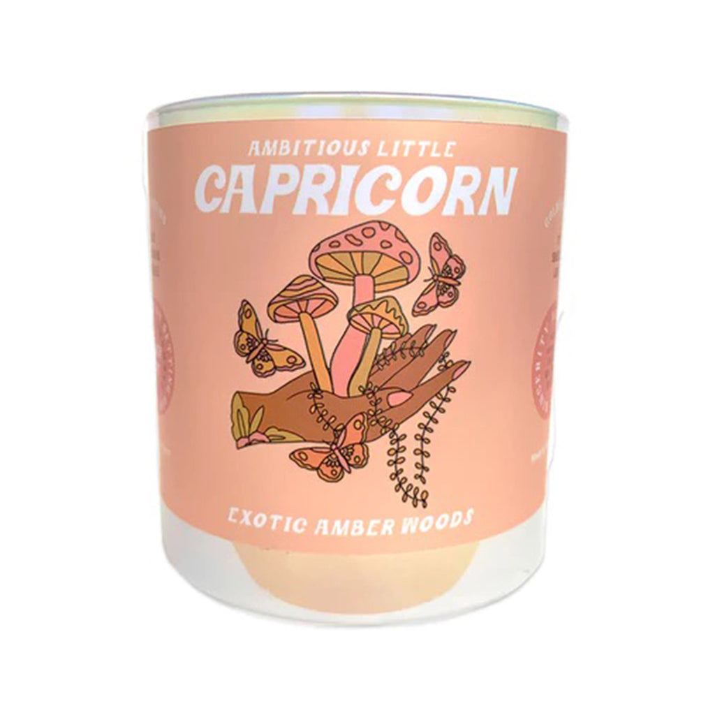 Golden Gems Ambitious Little Capricorn Exotic Amber Woods scented soy wax candle with illustrated peach label on an iridescent white glass tumbler, front view.
