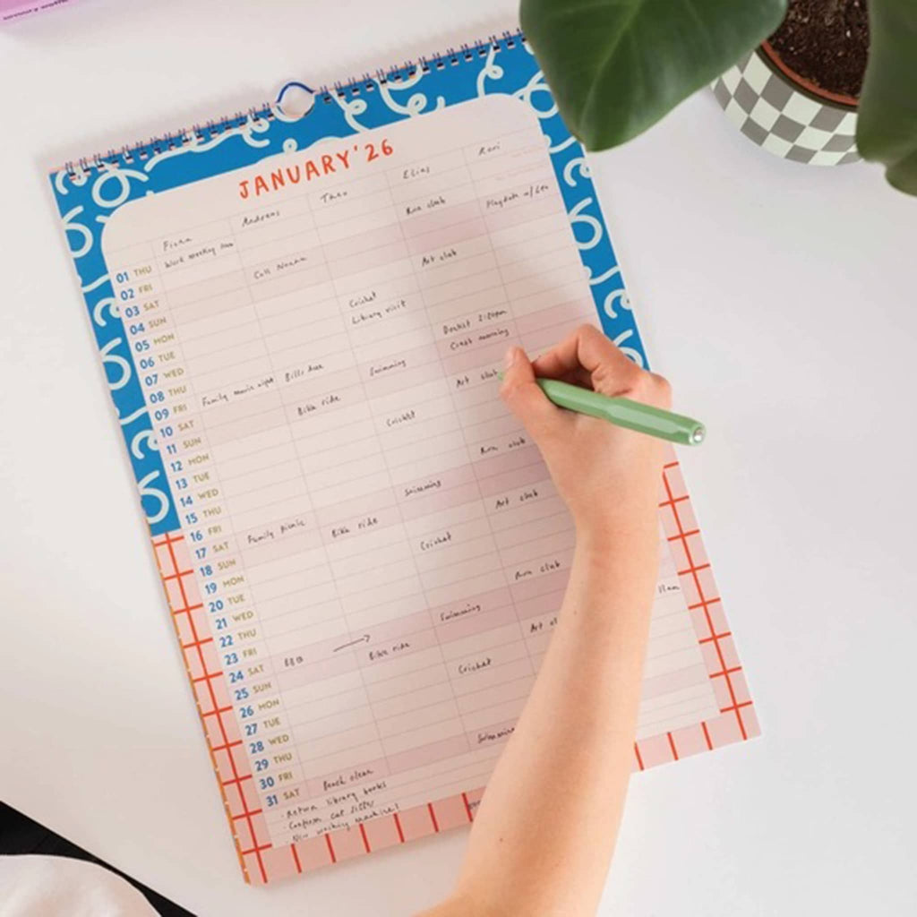 Person writing in a wall calendar with a hand holding a green pen on a white surface.