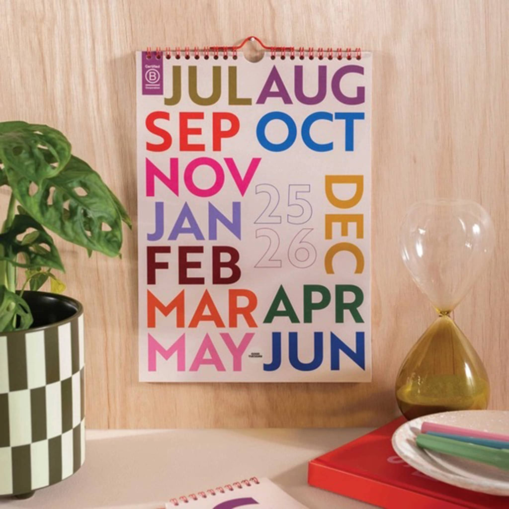Colorful academic year wall calendar on a wooden wall with a plant and hourglass in the foreground.
