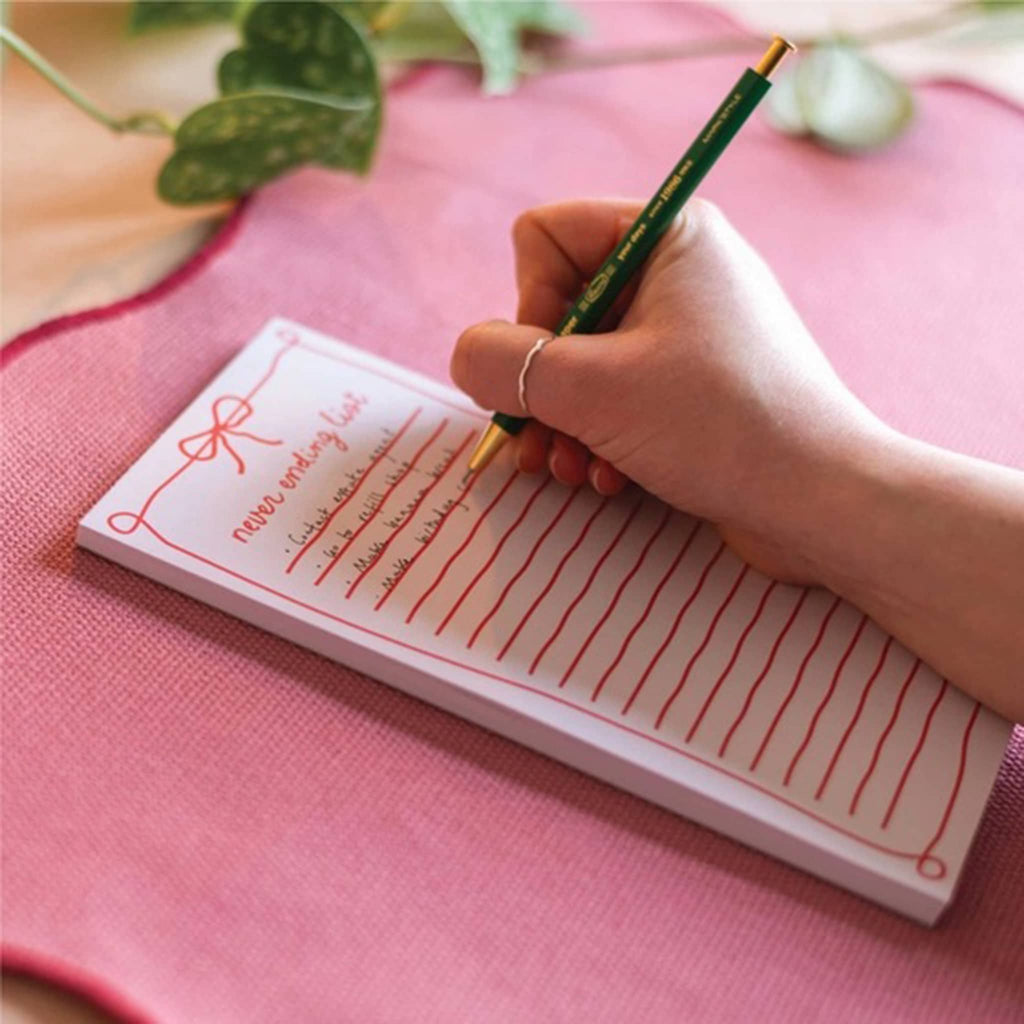 Hand writing on a lined notepad with a pen, placed on a pink surface.