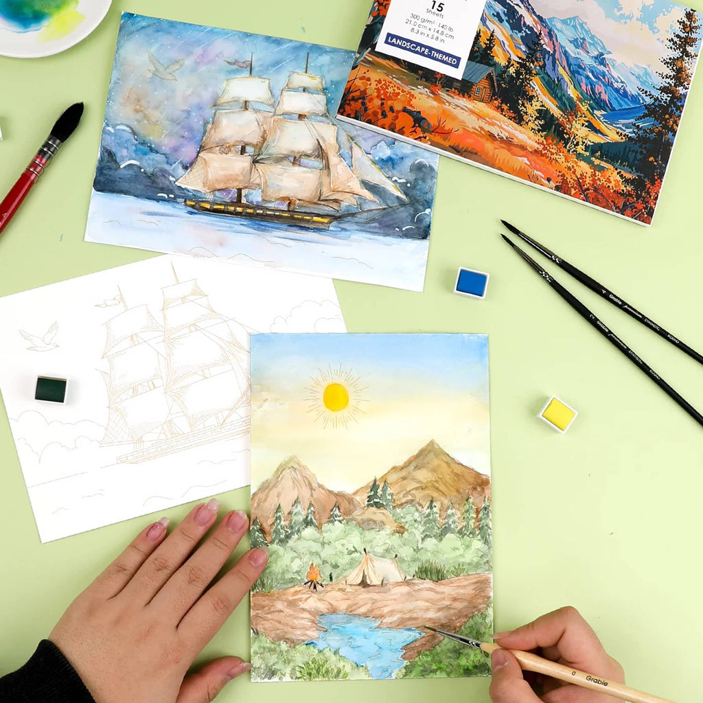 Artistic scene with painted cards featuring landscapes and a sailboat, with hands holding a paintbrush on a green surface.
