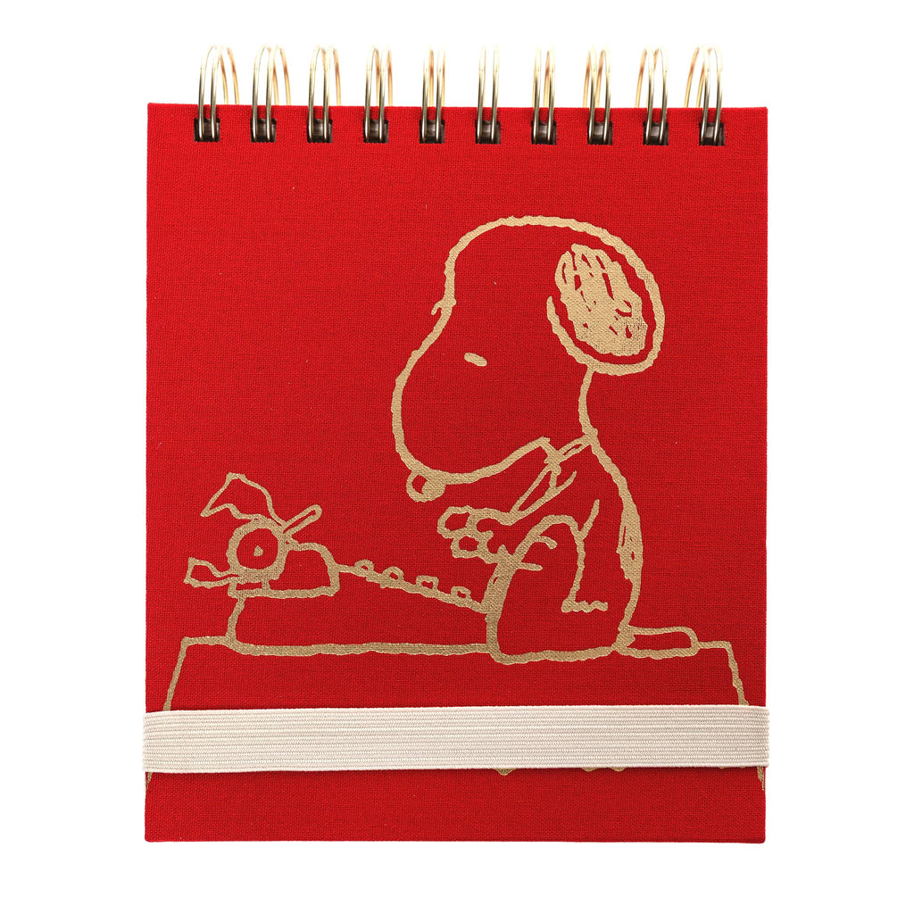 Red notebook with an illustration of Snoopy sitting on a desk with a typewriter in gold foil, on a white background.